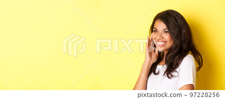 Close-up of beautiful african-american woman, smiling and looking left, gently touching face after applying makeup product, standing over yellow background Close-up of beautiful african-american woman, smiling and looking left, gently touching face after applying makeup product, standing over yellow background 97228256