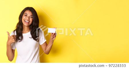 Portrait of good-looking african american woman in white t-shirt, showing thumbs-up and credit card, recommending bank, standing over yellow background 97228333