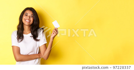 Portrait of beautiful african-american woman in white t-shirt, smiling pleased and showing credit card for shopping, standing over yellow background 97228337