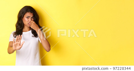 Young african-american woman in white t-shirt, shut her nose, looking disgusted and asking to stay away, dislike bad smell, standing over yellow background Young african-american woman in white t-shirt, shut her nose, looking disgusted and asking to stay away, dislike bad smell, standing over yellow background 97228344