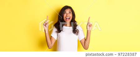 Portrait of amazed, happy african-american girl, smiling and pointing fingers up, standing over yellow background 97228387
