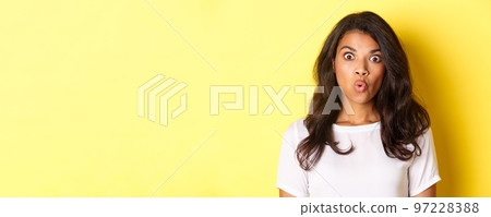 Close-up of amazed african-american girl, gasping and looking at something awesome, standing near copy space over yellow background Close-up of amazed african-american girl, gasping and looking at something awesome, standing near copy space over yellow background 97228388