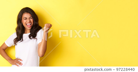Portrait of happy african-american woman, triumphing and winning, making fist pump and shouting for joy, standing pleased against yellow background 97228440