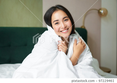 Cute and tender woman sitting in bed with warm duvet, smiling as looking out of the window, enjoying morning and comfort of blanket Cute and tender woman sitting in bed with warm duvet, smiling as looking out of the window, enjoying morning and comfort of blanket 97229461