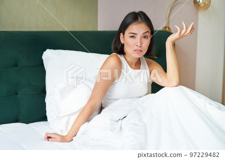 Portrait of puzzled asian woman lying in bed covered with warm duvet blanket, looking confused, shrugging with clueless face 97229482