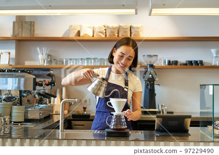 Smiling asian girl barista, pouring hot water from kettle, brewing filter coffee, standing behind counter in cafe Smiling asian girl barista, pouring hot water from kettle, brewing filter coffee, standing behind counter in cafe 97229490