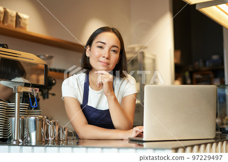 Portrait of smiling korean woman, barista in coffee shop, standing at counter with laptop, smiling and looking confident, self-employed female entrepreneur in her own coffee shop Portrait of smiling korean woman, barista in coffee shop, standing at counter with laptop, smiling and looking confident, self-employed female entrepreneur in her own coffee shop 97229497