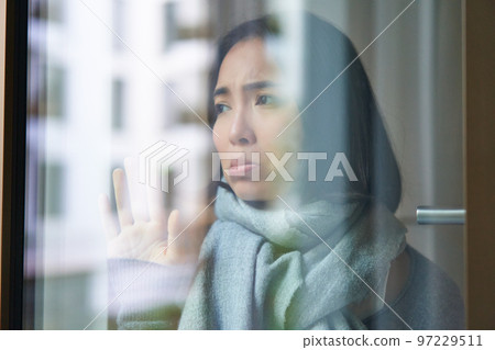 Portrait of sad ill korean woman looking out of the window, staying at home due to coronavirus, being sick and cant leave house Portrait of sad ill korean woman looking out of the window, staying at home due to coronavirus, being sick and cant leave house 97229511