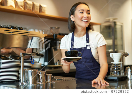Portrait of beautiful asian girl, student working part-time in cafe, holding cup of coffee, made order, looking for client, wearing uniform Portrait of beautiful asian girl, student working part-time in cafe, holding cup of coffee, made order, looking for client, wearing uniform 97229532