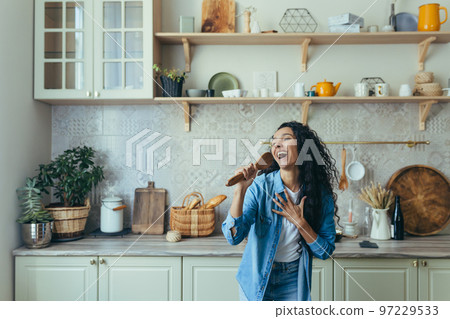 Young beautiful woman at home singing and dancing in the kitchen, cheerful Latin American woman having fun alone with curly hair. Young beautiful woman at home singing and dancing in the kitchen, cheerful Latin American woman having fun alone with curly hair. 97229533