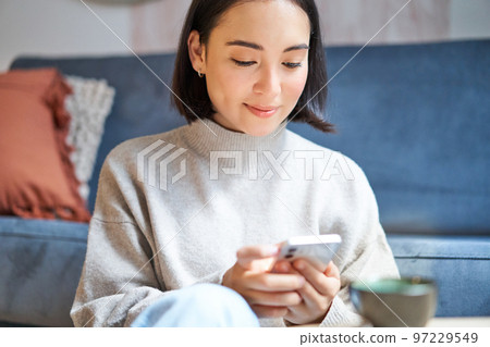 Portrait of smiling asian woman sits at home, uses smartphone app, orders delivery, messages someone on mobile phone application Portrait of smiling asian woman sits at home, uses smartphone app, orders delivery, messages someone on mobile phone application 97229549