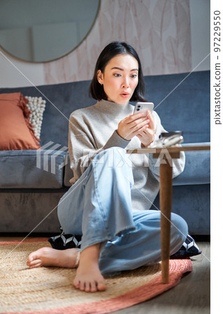 Vertical shot of young asian girl looks surprised at her mobile phone, reads amazing news, notification on smartphone app, sits at home on floor Vertical shot of young asian girl looks surprised at her mobile phone, reads amazing news, notification on smartphone app, sits at home on floor 97229550