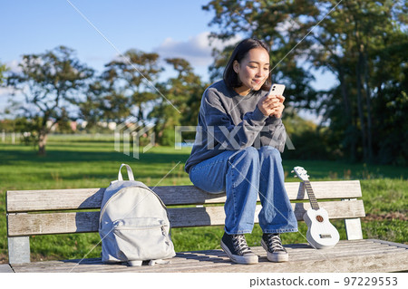 Young woman sitting in park on bench with ukulele, looking at smartphone, reading message on mobile phone and smiling Young woman sitting in park on bench with ukulele, looking at smartphone, reading message on mobile phone and smiling 97229553