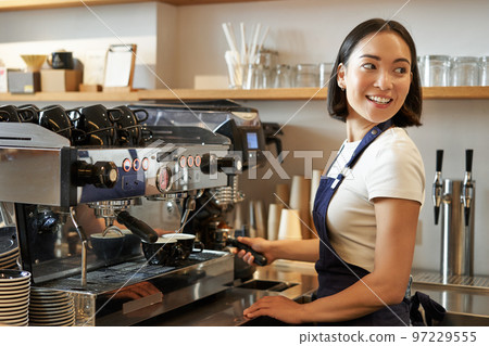 Smiling asian barista girl makes cappuccino with coffee machine, stands behind counter in cafe Smiling asian barista girl makes cappuccino with coffee machine, stands behind counter in cafe 97229555