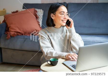 Portrait of smiling asian woman making phone call, working on laptop and having conversation on smartphone Portrait of smiling asian woman making phone call, working on laptop and having conversation on smartphone 97229573