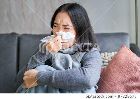 Close up of korean girl sneezing in napking, sitting at home in scarf, feeling unwell, concept of winter, heating cost and people Close up of korean girl sneezing in napking, sitting at home in scarf, feeling unwell, concept of winter, heating cost and people 97229575