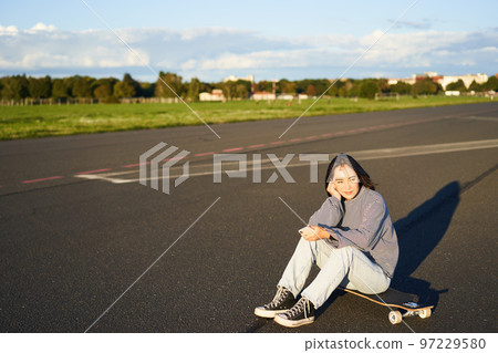 Skater girl sits on her skateboard on road, using smartphone, chatting on mobile app 97229580