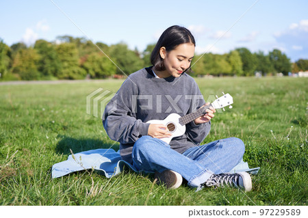 Beautiful asian girl sitting in park, playing ukulele and singing, relaxing outdoors on sunny spring day Beautiful asian girl sitting in park, playing ukulele and singing, relaxing outdoors on sunny spring day 97229589