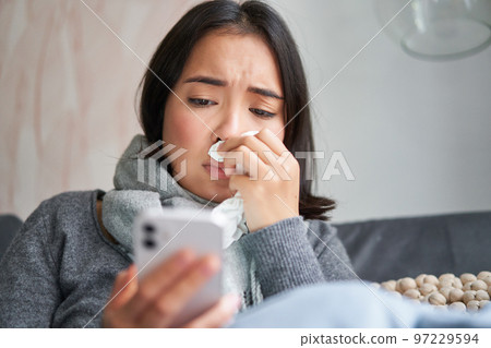 Ill korean woman sitting at home, sneezing, caught cold, reading news on mobile phone, freezing at her home because of heating problems 97229594