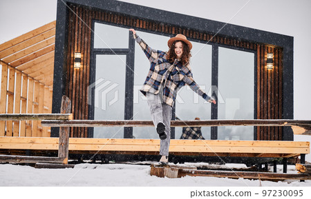 Curly young woman resting on terrace of modern barn house in the mountains. Happy female tourist enjoying winter holidays in new cottage. Curly young woman resting on terrace of modern barn house in the mountains. Happy female tourist enjoying winter holidays in new cottage. 97230095