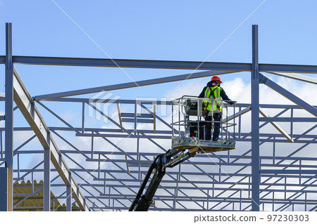 Worker in self propelled boom lift basket in uniform on a building steel framework background Worker in self propelled boom lift basket in uniform on a building steel framework background 97230303