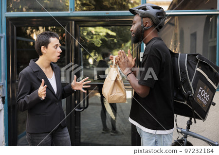 Woman shouting at office food delivery service courier, receiving wrong order, deliveryman carrying lunch late. Man giving impulsive unsatisfied customer takeaway meal near company building Woman shouting at office food delivery service courier, receiving wrong order, deliveryman carrying lunch late. Man giving impulsive unsatisfied customer takeaway meal near company building 97232184