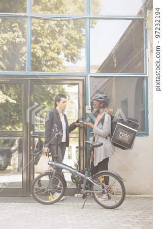 Customer paying for delivery, smartphone mobile wallet contactless payment, nfc technology. African american courier holding pos terminal, standing near office building outdoors 97232194