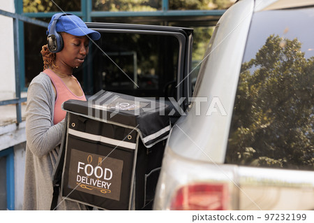 African american courier in headphones delivering takeaway food by car, waiting for customer near office building outdoors. Delivery service worker taking out backpack from automobile 97232199