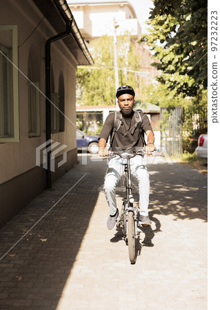 Food delivery courier riding bicycle outdoors, delivering restaurant meal on sunny day portrait. Deliveryman with customer order in backpack box, looking at camera front view Food delivery courier riding bicycle outdoors, delivering restaurant meal on sunny day portrait. Deliveryman with customer order in backpack box, looking at camera front view 97232223