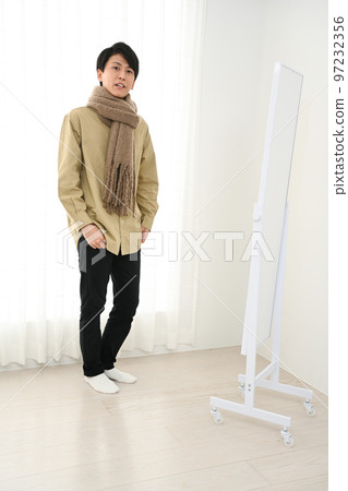 A young man wearing a scarf while looking at a full-length mirror indoors A young man wearing a scarf while looking at a full-length mirror indoors 97232356