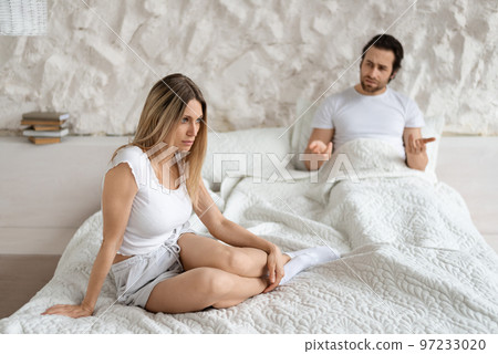 Unhappy angry husband freaking out and yelling at sad wife, sitting on bed in bedroom interior 97233020