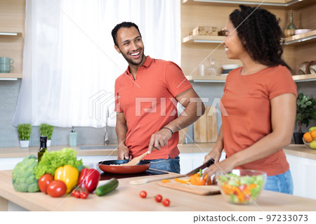 Smiling young black husband and wife in red t-shirts cuts salad, prepares lunch, cooking together in kitchen 97233073