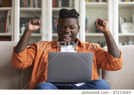 Emotional black man with laptop celebrating success, home interior Emotional black man with laptop celebrating success, home interior 97233084