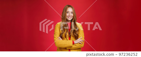 Charming successful young female in yellow autumn coat smiling broadly holding hands crossed on body in confident self-assured pose standing delighted and happy over red background 97236252
