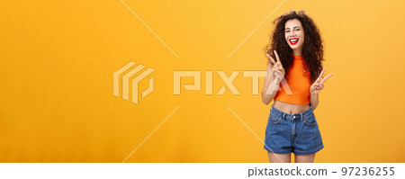 Outgoing friendly-looking attractive young female with curly haircut in cropped top and shorts showing peace or victory signs with both hands and smiling cheerfully having fun over orange background Outgoing friendly-looking attractive young female with curly haircut in cropped top and shorts showing peace or victory signs with both hands and smiling cheerfully having fun over orange background 97236255