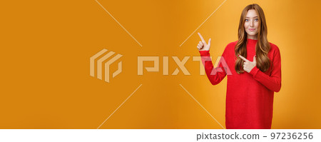 Soft and gentle gorgeous ginger girl with freckles in red warm sweater smiling joyfully pointing at upper left corner smiling showing curious place, suggesting visit it over orange background Soft and gentle gorgeous ginger girl with freckles in red warm sweater smiling joyfully pointing at upper left corner smiling showing curious place, suggesting visit it over orange background 97236256