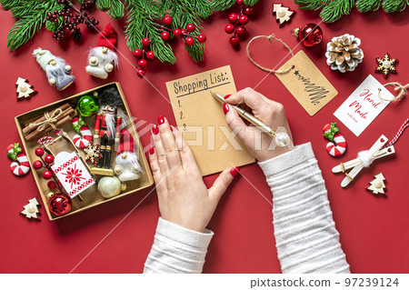 female hand holds pen and writing Christmas shopping list, gift ideas on white notepad on red background Top view Flat lay Holiday card Woman making Christmas presents list, Planning, buying concept. female hand holds pen and writing Christmas shopping list, gift ideas on white notepad on red background Top view Flat lay Holiday card Woman making Christmas presents list, Planning, buying concept. 97239124