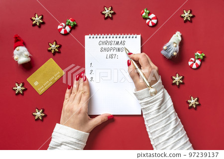 female hand holds pen and writing Christmas shopping list, gift ideas on white notepad on red background Top view Flat lay Holiday card Woman making Christmas presents list, Planning, buying concept. 97239137