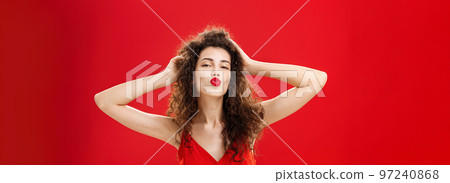 Waist-up shot of confident flirty and elegant young rich woman with curly hairstyle in red lipstic and evening dress touching hair folding lips in mwah giving kiss to admirer feeling self-assured Waist-up shot of confident flirty and elegant young rich woman with curly hairstyle in red lipstic and evening dress touching hair folding lips in mwah giving kiss to admirer feeling self-assured 97240868
