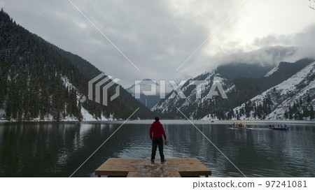 The guy standing on the pier admires mountain lake The guy standing on the pier admires mountain lake 97241081