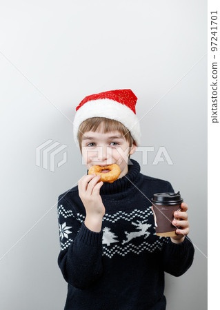 A blond boy in a Santa hat, mittens and a warm winter sweater, chewing a donut. Light background, vertical, copy of the space 97241701