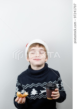 A boy in a Santa hat and a warm sweater with a donut and a drink in his hands, Eating with his eyes closed, smiling with pleasure A boy in a Santa hat and a warm sweater with a donut and a drink in his hands, Eating with his eyes closed, smiling with pleasure 97241707