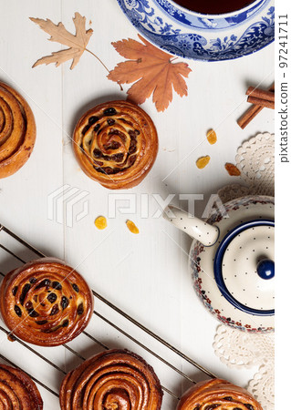 Buns with cinnamon and raisins cool on the grill under a lace napkin, on a light background. With a teapot, autumn leaves and a cinnamon stick. Vertical view 97241711