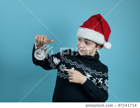 A boy in a warm sweater and a New Year's hat with a vintage alarm clock in his hands. Portrait on a blue background. New Year's layout for text A boy in a warm sweater and a New Year's hat with a vintage alarm clock in his hands. Portrait on a blue background. New Year's layout for text 97241714
