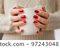 Woman's hands with red manicure and gold foil on the nails. Trendy autumn and winter nail design. Woman with a beautiful manicure holding a white knitted cup. The concept of cozy Christmas holidays. 97243048