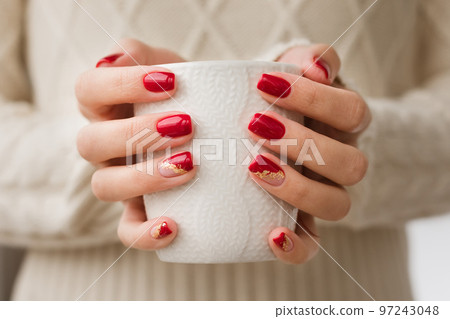 Woman's hands with red manicure and gold foil on the nails. Trendy autumn and winter nail design. Woman with a beautiful manicure holding a white knitted cup. The concept of cozy Christmas holidays. 97243048