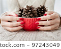 Woman's hands with red manicure and gold foil on the nails. Trendy autumn and winter nail design. Woman with a beautiful manicure holding a red knitted cup. The concept of cozy Christmas holidays. 97243049