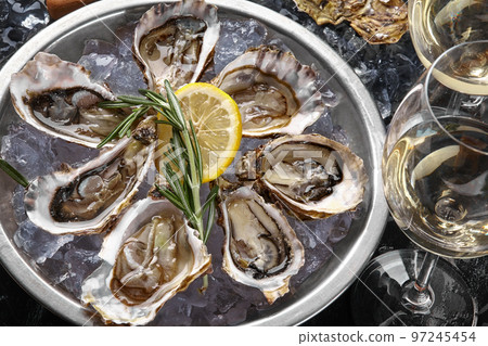 Fresh opened oysters, ice, lemon on a round metal plate and champagne are on a black stone textured background. Top view. Close-up shot. 97245454