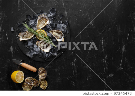 Fresh opened oysters, ice and lemon on a round slate, black stone textured background. Top view with copy space. Close-up shot. 97245473