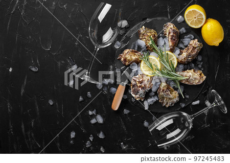 Fresh closed oysters, ice, lemon on a round slate and two empty glasses are on a black stone textured background. Top view with copy space. Close-up. Fresh closed oysters, ice, lemon on a round slate and two empty glasses are on a black stone textured background. Top view with copy space. Close-up. 97245483